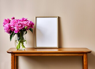 Empty picture frame mockup on beige wall. Wooden table. Glass vase with pink peonies bouquet. Elegant working space, home office concept. Interior design.