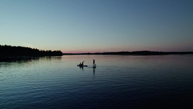 Aerial orbit shot, woman with kids on stand up paddle board on calm lake with water ripples in evening twilight. Silhouettes of the woods and orange purple blue sunset sky in the background