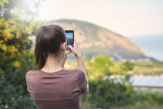 back view of young woman photographing the mountain landscape by smartphone camera at the sunset
