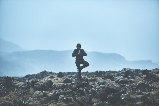 Woman in waterproof clothing stands in yoga pose on volcanic Lava fields on background of mountains of Iceland.
