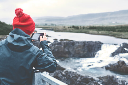 a woman tourist in waterproof clothing and red knit hat photographing a small river waterfall in Iceland under the rain