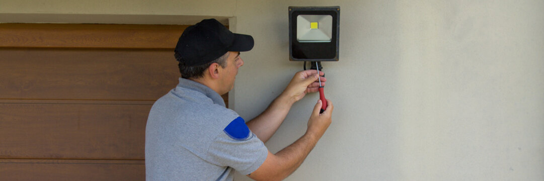 Image of an electrician on a scissor ladder using his tools to install a spotlight on a house. Illumination of the darkest parts of the house. Horizontal banner 
