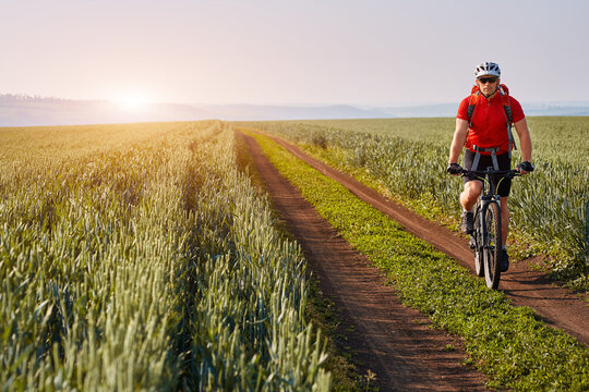 Young cyclist with mountain bicyclist on the path of the field in the countryside against sunrise. Sportsman dressed in the sportwear, with helmet, sunglasses and red backpack. Beautiful landscape lik