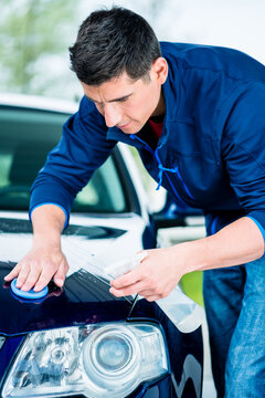 Happy young man looking at camera while waxing a blue car outdoors at car wash