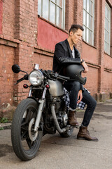 Fototapeta na wymiar Handsome rider biker guy in black leather jacket, boots and style denim sit on classic style cafe racer motorcycle. Bike custom made in vintage garage. Brutal fun urban lifestyle. Outdoor portrait.