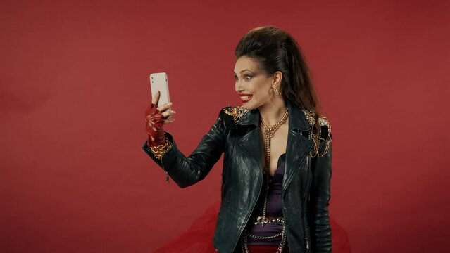 Woman speaks on video call on smartphone on red background. A woman is embarrassed, flirts, waves her hand, sends an air kiss. Having finished the conversation, he puts the phone to his chest.