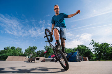A subcultural man from generation x is balancing on one wheel on his bmx in a skate park.