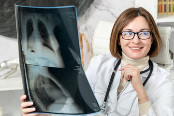 Portrait of young female doctor pulmonologist sitting at the desktop in modern office and holding x...