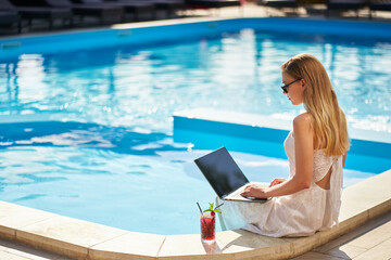 Self employed woman doing remote work with laptop sitting near swimming pool at tropical spa with...