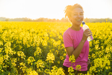 Fototapeta premium Outdoor portrait of beautiful happy mixed race African American girl teenager female young woman athlete runner drinking water from a bottle in a field of yellow flowers at sunset in golden evening su