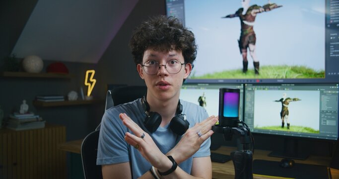 Young 3D designer speaks on camera using microphone, records video tutorial for blog about video game design and 3D modeling at home studio. Software interface on computer and big digital screen.