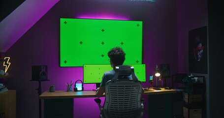 Young editor, 3D designer works remotely at home studio, edits movie, develops video game, creates 3D animation. Teenager sits in front of multi-monitor computer and big digital screen with chromakey.