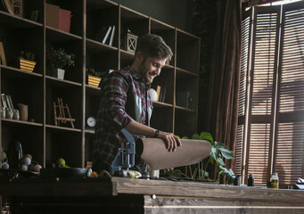 Work with smile on face. Young man leather worker holding in hand folded roll of brown leather over wooden working table. Handmade product from natural leather