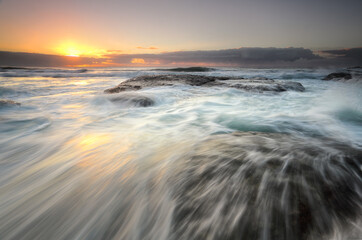 Ocean flowing rapidly over exposed rocks as the sun's glow peeks from behind clouds on the horizon.  Location, Bungan Beach, nortnern beaches Sydney Australia