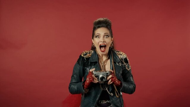 A woman passes with a vintage camera, stops abruptly and takes a lot of shots on the camera. The woman joyfully jumps in place and runs away. Woman with a camera in the studio on a red background.