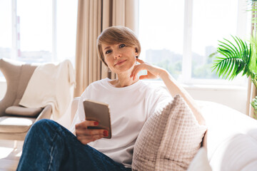 Middle aged woman with smartphone sitting on sofa in living room