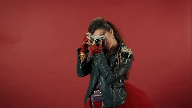 A woman sets up an old camera, points the lens and takes pictures. Woman freak, with bright makeup and bouffant in the studio on a red background. Advertising.