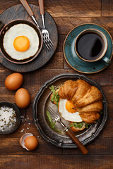Croissant sandwich with fried egg and arugula, cup of coffee, fried egg in pan, salt and eggshell on wooden background. Flat composition. Delicious and healthy breakfast