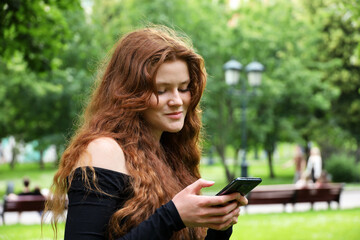 Smiling girl with long red hair and freckles using mobile phone standing in summer park. Concept of flirting and online dating