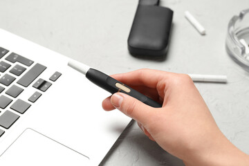 Woman holding modern electronic cigar near laptop on grey background
