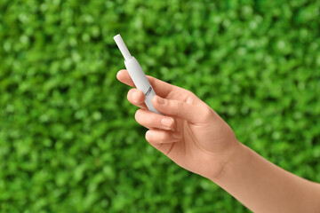 Female hand holding modern electronic cigar with stick against green nature background