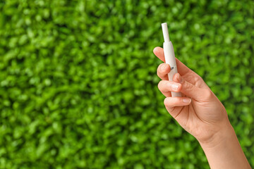 Female hand holding modern electronic cigar with stick against green nature background