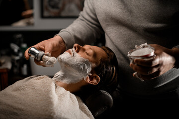 Barber applying shaving cream on client's beard before shaving in barber shop