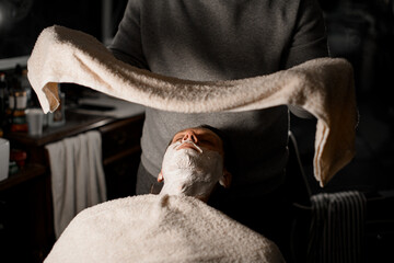 Hands of barber applying hot towel to male client's face to moisturize beard hairs before shaving