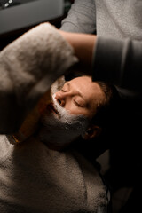 Hands of barber applying hot towel to male client's face to moisturize beard hairs before shaving process