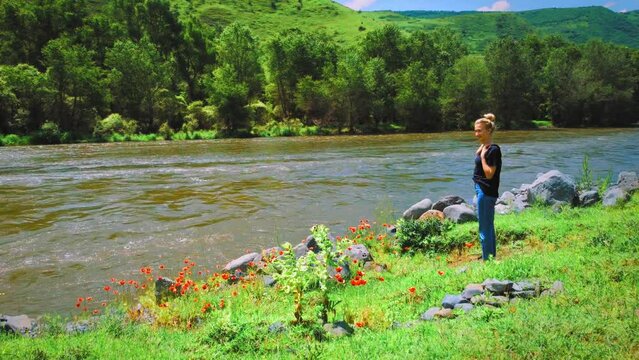 Young woman in jeans and a T-shirt stands near a mountain river against the background of mountains.Beautiful landscape with a river, trees,poppies and a girl from the back.Concept of travel,lifestyle