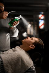 Professional hairdresser sprinkles green liquid from vintage desidned stylish bottle above client's face in modern barbershop