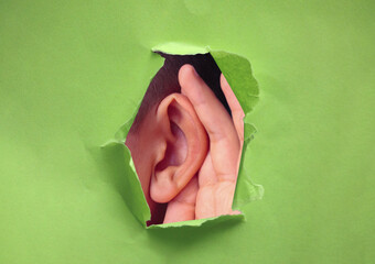 Close up ear and hand through a torn hole in green paper background, free copy space, closeup. The...