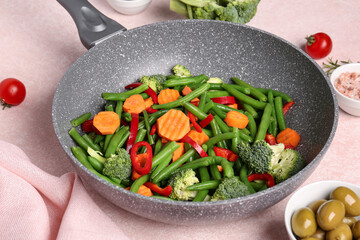 Frying pan with fresh vegetables on pink background