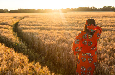 African woman in traditional clothes standing, looking to the sun, hand to eyes, in field of barley...