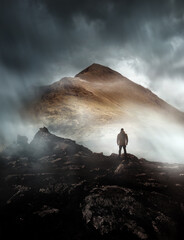 A person hiking looks onwards at a mountain shrouded in mist and clouds with the peak visible. Scenic landscape photo composite.