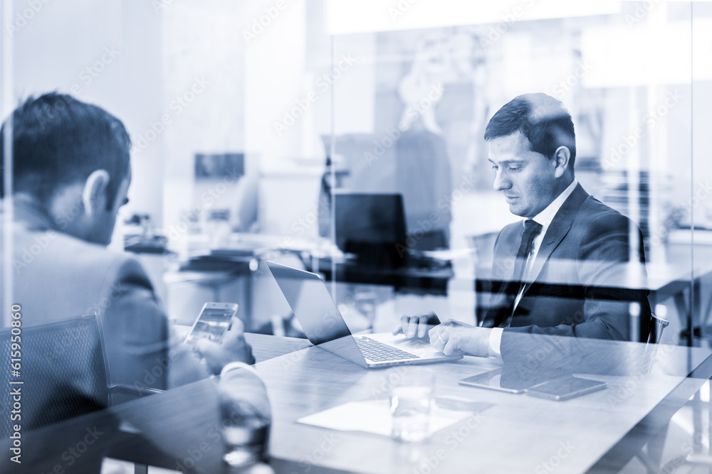 Poster through the window view of two young businessmen using laptop computer and smart phone devices at wo