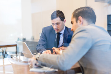 Through the window view of two young businessmen using laptop computer at business meeting in corporative office.