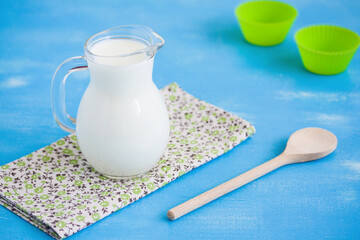 Milk in glass jug on green and brown floral pattern napkin, wooden spoon and green baking dish on blue wooden  background. Focus on wooden spoon and napkin.