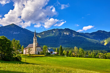 Fototapeta premium Austria. Traditional church with chapel in village among picturesque landscapes in austrian Alps mountains. Summer green lawns and fields. Knolls covered with forests and trees. Blue sky with clouds.
