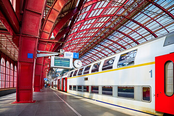 Antwerp, Belgium. Central indoor railway station. Platform made of red metal constructions with clock and panel with departure or arrival schedule. Modern double decker high-speed train - obrazy, fototapety, plakaty