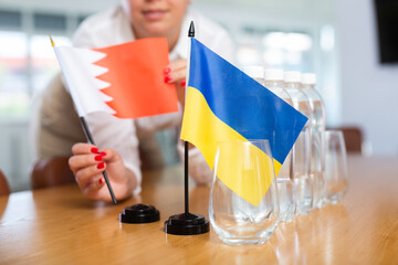 Female office administrator placing Kingdom of Bahrain flag on table near national flag of Ukraine,...