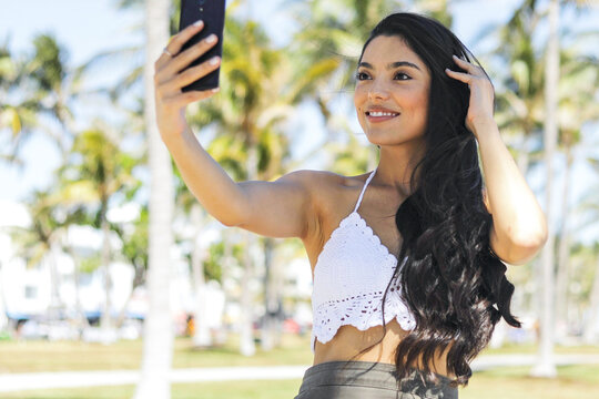 Content ethnic model with long shiny hair posing sensually at camera of smartphone while taking selfie.