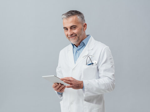 Confident mature doctor connecting with a digital touch screen tablet and using apps, online medical diagnosis and consultation concept