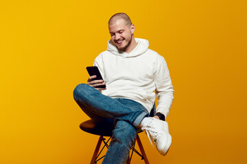 Excited guy sitting on chair, using mobile application on modern smartphone, chatting with friends or watching funny video, yellow background