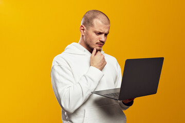 Portrait of a doubtful bearded man in white hoodie holding laptop computer while touching chin with...