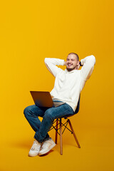 Fototapeta na wymiar Vertical photo of happy man sitting on chair using laptop computer, holding hands behind neck, rest, relax, isolated over yellow background
