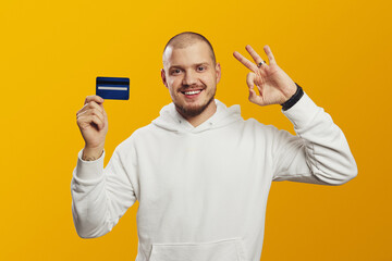 Excited young caucasian man in white hoodie holding credit bank card while showing OK gesture,...
