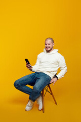 Smiling young man using smartphone while sitting on chair isolated on yellow background. Excited...