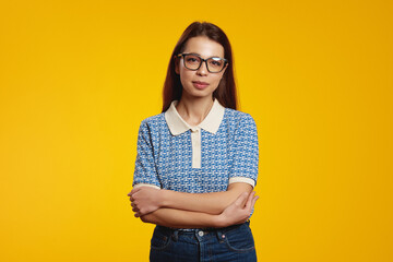 Serious young girl wearing blue shirt and eyeglasses standing with crossed arms against yellow background