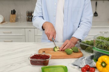 Man cutting fresh broccoli with knife near containers at white marble table in kitchen, closeup and space for text. Food storage